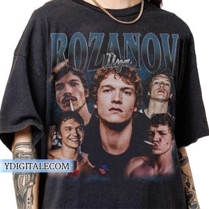 May include: Dark gray t-shirt featuring a collage of images of a person with the name "ROZANOV" and "Ilya" printed in a blue font. The images show the person in various poses, some with a cigarette.