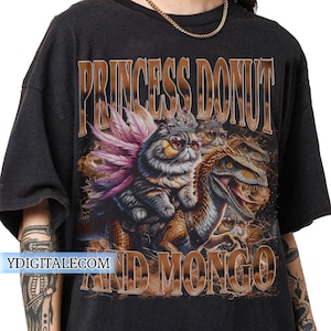 May include: Black t-shirt with a graphic featuring a cat wearing sunglasses and a crown, riding a dinosaur. The text "PRINCESS DONUT AND MONGO" is printed above and below the image in a brown, textured font.