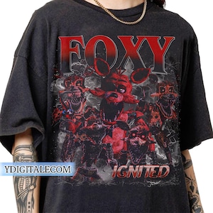Könnte beinhalten: Dunkelgraues T-Shirt mit einer roten Grafik der Figur Foxy aus dem Videospiel Five Nights at Freddy's. Das Wort "FOXY" ist in großen roten Buchstaben über der Figur aufgedruckt, darunter das Wort "IGNITED".