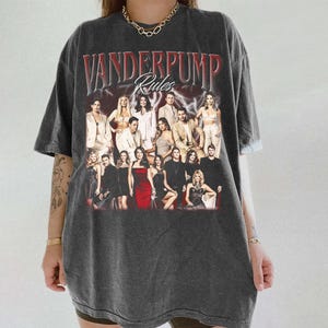 May include: Dark gray oversized t-shirt featuring the text "Vanderpump Rules" in red and white, with a group photo of the cast. The shirt has a vintage, worn look.