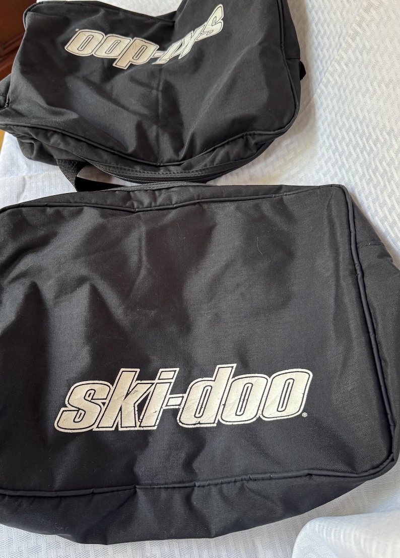 May include: Two black Ski-Doo branded bags. The larger bag has the words "ski-doo" in white block letters. The smaller bag has the words "doo-ski" in white block letters. Both bags are made of a durable material.