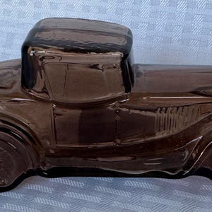 May include: A brown glass antique car-shaped bottle with a black screw-on cap. The vintage vehicle has detailed features, including fenders, wheels, and a cab. The bottle is likely a decorative item or a container for liquids.