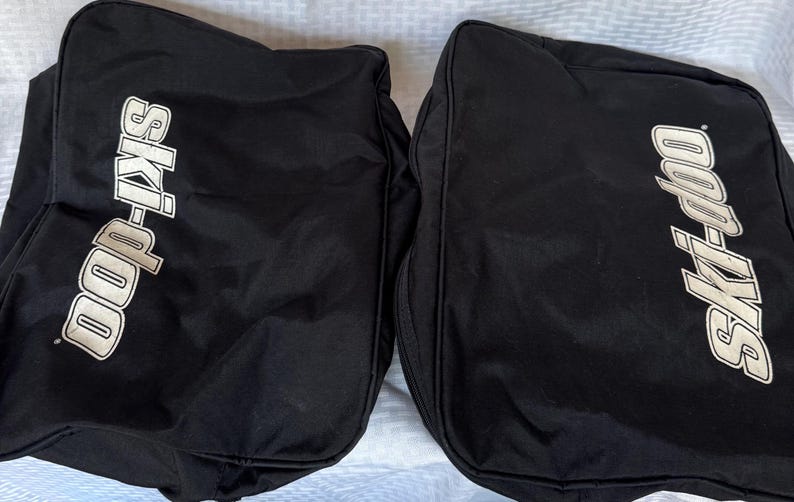May include: Two black Ski-Doo branded bags. Each bag has the Ski-Doo logo in white, with the brand name in a stylized font. The bags appear to be made of a durable, possibly water-resistant material, suitable for outdoor gear.