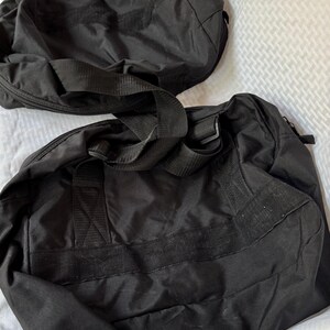 May include: Two black duffel bags with sturdy handles and shoulder straps. The bags are made of a durable, possibly water-resistant material. The bags are designed for travel or carrying sports equipment.