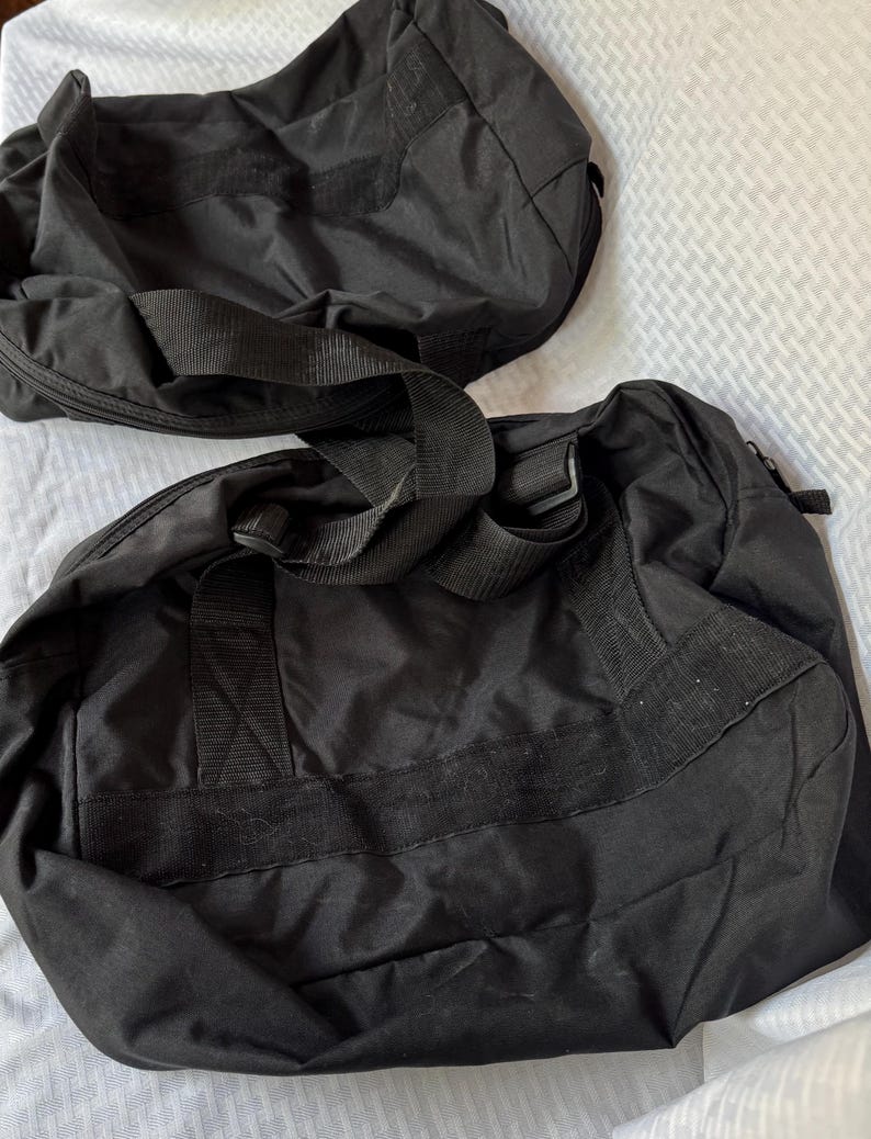May include: Two black duffel bags with sturdy handles and shoulder straps. The bags are made of a durable, possibly water-resistant material. The bags are designed for travel or carrying sports equipment.