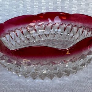 May include: A clear glass bowl with a ruby red rim and a diamond-cut pattern. The bowl has a wavy edge and is likely used for serving or display. The bowl is on a white surface.