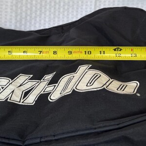 May include: Black snowmobile cover with the "Ski-doo" logo in white. The cover measures approximately 16 inches in length, as measured by a yellow tape measure.
