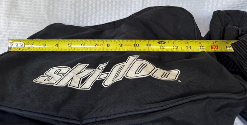May include: Black snowmobile cover with the "Ski-doo" logo in white. The cover measures approximately 16 inches in length, as measured by a yellow tape measure.