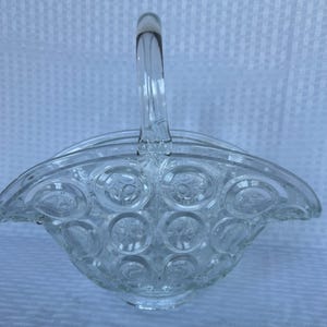 May include: Clear glass basket with a handle. The basket features a pattern of circles with a starburst design in the center. The handle is arched and attached to the rim of the basket.