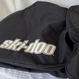 May include: Black snowmobile bag with the white "Ski-doo" logo. The bag appears to be made of a durable, water-resistant material. The bag is designed to protect snowmobile components during storage or transport.