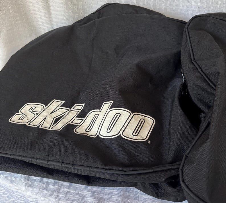 May include: Black snowmobile bag with the white "Ski-doo" logo. The bag appears to be made of a durable, water-resistant material. The bag is designed to protect snowmobile components during storage or transport.