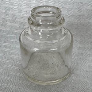 May include: A clear, cylindrical glass inkwell with a rounded base and a wide, slightly flared opening. The inkwell has a short neck and a smooth, transparent surface. The inkwell is sitting on a patterned, light-colored surface.