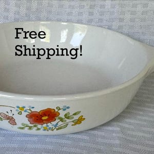 May include: A white ceramic skillet-shaped dish with a handle, featuring a colorful floral pattern on the exterior side. The design includes red, yellow, and blue flowers with green leaves and a small butterfly. The interior is plain white.