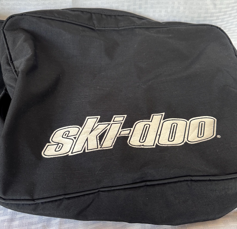 May include: Black bag with the white text "ski-doo" in a stylized font. The bag appears to be made of a durable material and is likely designed for carrying equipment. The bag is rectangular and has a simple design.