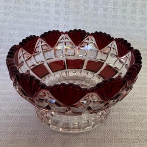 May include: A clear and deep red glass bowl featuring a cut crystal pattern with a scalloped rim. The bowl has a faceted geometric design throughout the body, alternating between transparent clear glass and solid dark red glass sections.