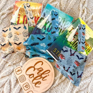 May include: A collection of handmade bookmarks and book sleeves with various designs. The bookmarks feature tassels and themed patterns with bats, cars, and text. The book sleeves have coordinating patterns. A wooden sign with social media icons is also present.