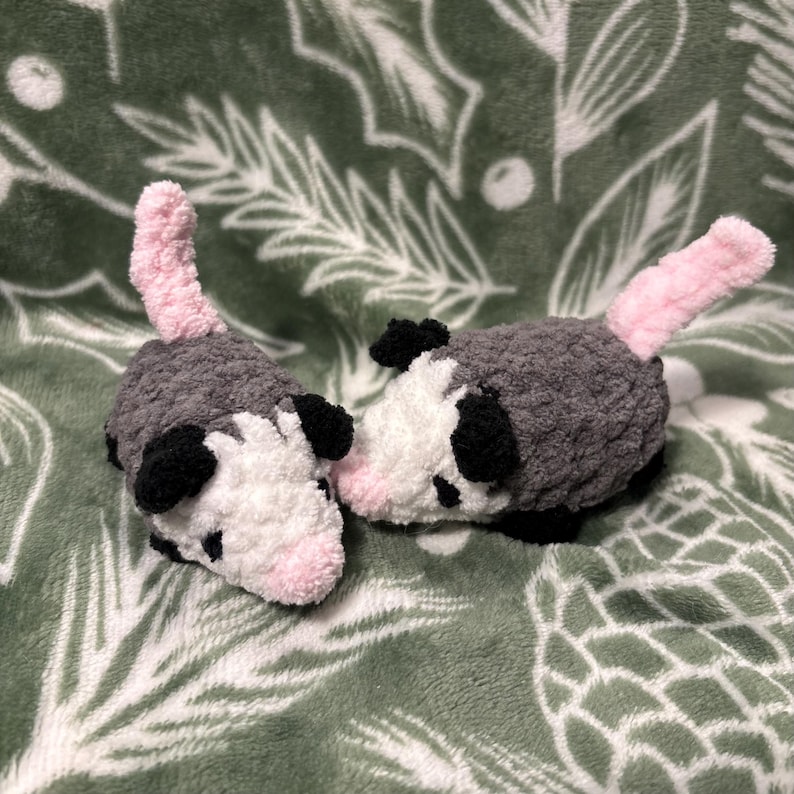 May include: Two handmade crochet opossums. Each has a gray body, white face, black ears, and a pink tail. They are lying on a green blanket with a white leaf pattern. The opossums are soft and plush.