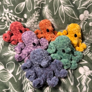 May include: A collection of six plush octopuses in various colors: red, orange, purple, blue, green, and yellow. Each octopus has a black eye and is made of a soft, textured material. The octopuses are arranged on a green and white patterned blanket.