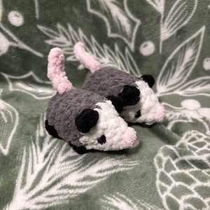 May include: Two handmade crochet opossums. The plush toys are gray with white faces, black ears, and pink noses and tails. The opossums are lying on a green blanket with a white leaf pattern.