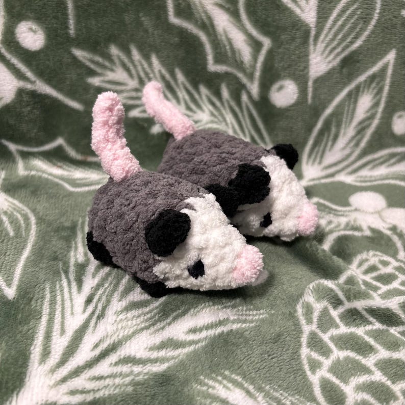 May include: Two handmade crochet opossums. The plush toys are gray with white faces, black ears, and pink noses and tails. The opossums are lying on a green blanket with a white leaf pattern.