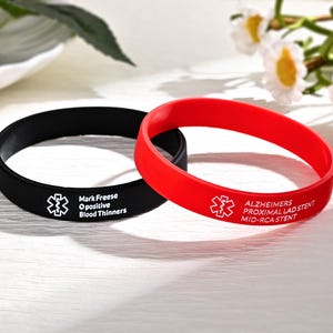 May include: Two medical alert bracelets. One is black with white text: "Mark Freese O positive Blood Thinners" and a medical symbol. The other is red with white text: "Alzheimers Proximal LAD Stent Mid-RCA Stent" and a medical symbol.