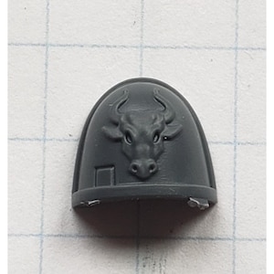 May include: Gray plastic miniature of a space marine shoulder pad with a bull's head emblem.