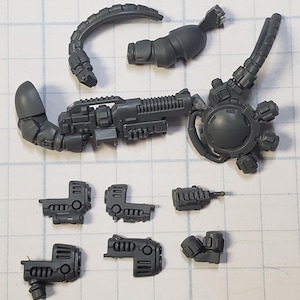 May include: A set of gray plastic model pieces for a sci-fi robot or miniature figure. The pieces include a gun, a head, a torso, and several arms.