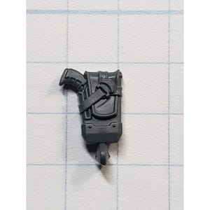 May include: A dark gray miniature accessory, possibly for a model or game. It features a rectangular pouch with a flap and strap, and a small, attached object resembling a gun. The item has a hook at the bottom.