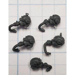 May include: Four dark gray miniature gas mask helmets with curved breathing tubes. The helmets have detailed features, including circular and rectangular elements. The pieces are arranged on a white grid background.