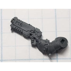 May include: A gray plastic miniature weapon, possibly a pistol, with a detailed design. The weapon is held by a miniature arm with a square socket at the end. The item is likely a component for a tabletop game or model.