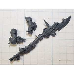 May include: A gray plastic miniature weapon with ornate details, including a large blade, spikes, and decorative elements. Two armor pieces are also included. The components are designed for a tabletop game.
