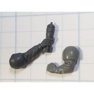 May include: Two gray plastic miniature arms for a model figure. One arm is holding a small gray plastic grenade. The other arm is bent at the elbow.