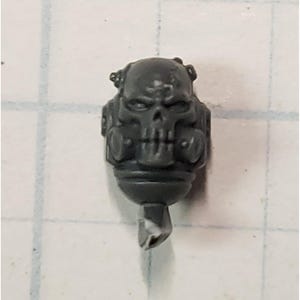 May include: A gray plastic miniature head featuring a skull design with a gas mask. The skull has detailed facial features, including eye sockets, a nose, and teeth. The miniature has a cylindrical base with a broken tip.