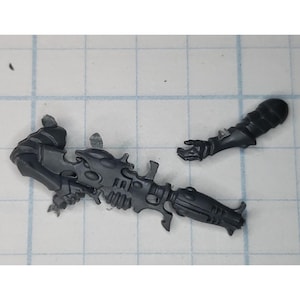May include: A dark gray plastic miniature weapon and arm, likely for a tabletop game. The weapon has a complex, futuristic design with multiple barrels and intricate details. The arm is detached, with armored segments and a detailed hand.