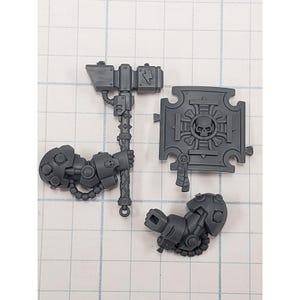 May include: A collection of unpainted, gray plastic miniature wargaming components. Includes a large hammer with a lightning bolt design, a shield with a skull emblem, and two mechanical arm pieces. The pieces are designed for assembly.