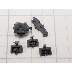 May include: A collection of gray plastic model kit parts, including a rectangular piece with a circular element, a cylindrical piece, and several other geometric shapes. The parts are arranged on a grid-lined surface.