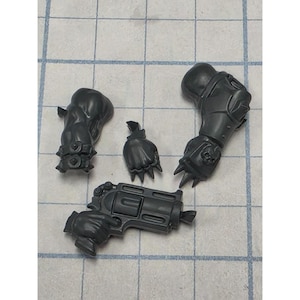 May include: A set of four unpainted dark gray plastic miniature model parts including a gloved hand holding a revolver, a separate clenched fist, a forearm piece with a spiked cuff, and a larger armored arm piece featuring a skull emblem.
