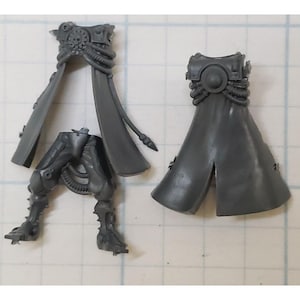 May include: Two unpainted gray plastic miniature model components. One piece features a torso with a long coat and a skull emblem, connected to separate legs. The second piece is a separate torso with a long coat and a circular chest plate.