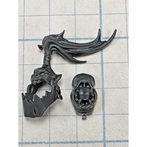 May include: A dark gray plastic miniature component featuring a jagged, trap-like jaw piece with a flowing, swept-back crest attached to the top. Beside it lies a separate, smaller dark gray plastic piece showing a detailed open mouth with teeth.
