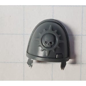 May include: Gray plastic miniature shoulder pad with a skull and starburst design.