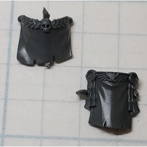 May include: Two gray plastic miniature banners for a tabletop wargame. One banner features a skull and crossbones, the other features a winged eagle.