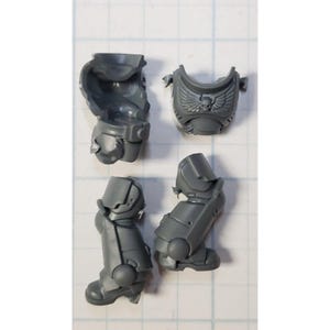 May include: A collection of unpainted, gray plastic miniature parts, including two leg pieces, a chest piece with a winged skull emblem, and an arm piece. These components appear to be from a model kit, likely for tabletop gaming or hobby projects.