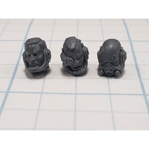 May include: Three gray miniature sci-fi heads with detailed features. Each head has a unique design, including helmets, visors, and facial expressions. The heads are likely for tabletop gaming or model building.