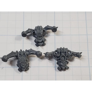 May include: Three gray plastic miniature model components. Each component features a central body with attached arm-like structures and intricate mechanical detailing. The pieces are designed for a tabletop game or hobby project.