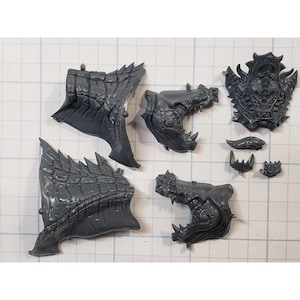 May include: A collection of unpainted, gray plastic miniature parts, including dragon-like wings, jaws with teeth, and a shield. The pieces are designed for a tabletop game or hobby project, with intricate details and textures.