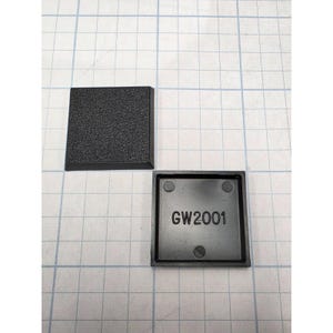 May include: Two square black plastic end caps with a textured top surface. One cap is positioned face up showing the grain, while the other is flipped over to reveal a smooth underside with the molded text GW2001 and four small circular indentations.