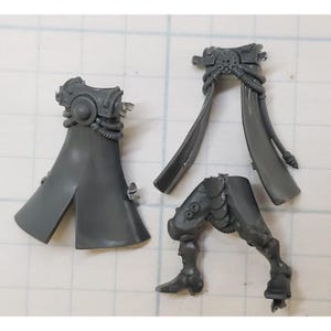 May include: Three gray plastic miniature components, likely for a model kit. The pieces include a cape-like structure, a flowing garment, and a leg with armor details. The parts are unpainted and show intricate molded designs.