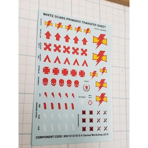 May include: A sheet of White Scars Primaris transfer decals. The sheet features red and yellow lightning bolts, arrows, skulls, and other symbols. Text includes "WHITE SCARS PRIMARIS TRANSFER SHEET" and "Games Workshop 2019".