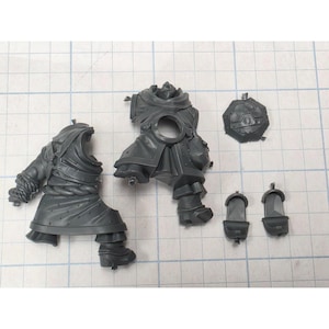 May include: A set of unpainted grey plastic miniature model components including two torso sections with draped robes and rope belts, a circular shield-like piece, and two small boot or foot attachments, all arranged on a grid surface.