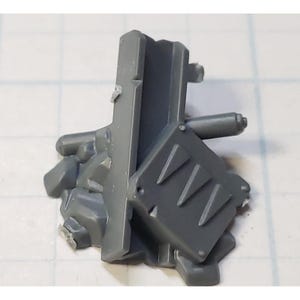 May include: A gray plastic miniature terrain piece, possibly for a tabletop game. It features a rectangular structure with a textured surface, a small angled shield with three triangular cutouts, and a cylindrical element.